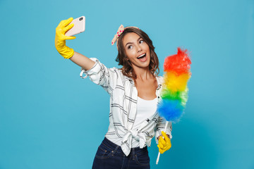 Cute housewife 20s in yellow rubber gloves for hands protection taking selfie on mobile phone while holding colorful duster brush during cleaning, isolated over blue background