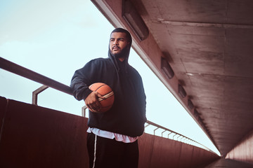 Dark-skinned bearded guy dressed in a black hoodie and sports shorts leaning on a guardrail while...