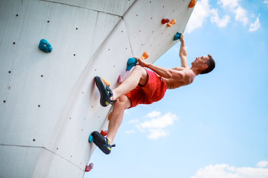 Photo from bottom of strong athletic man exercising on wall for climbing against blue sky with clouds