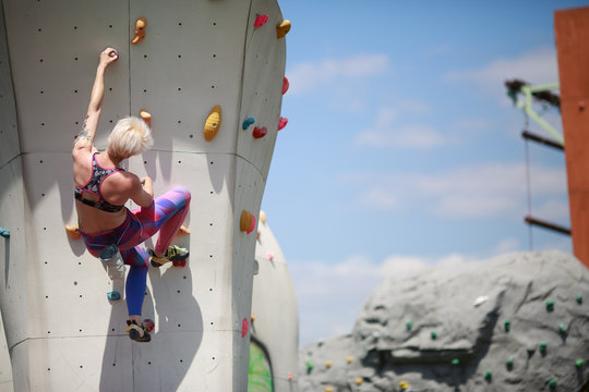 Photo from back of young blonde with short haircut in sports clothes practicing on wall for rock climbing against blue sky