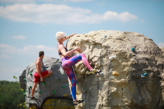 Photo of sports man and blonde sportswoman in sunglasses climbing on boulders for rock climbing against blue sky with clouds
