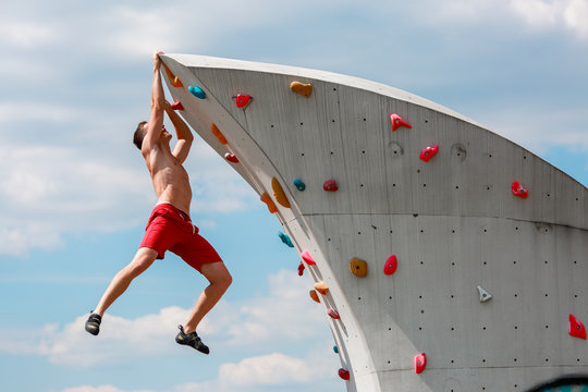 Photo of young sporty brunet in red shorts hanging on wall for rock climbing against blue sky with clouds