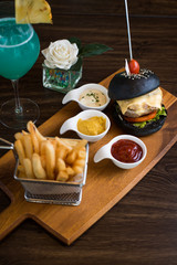 Black burger on board on dark wooden table background. Black burger with meat patty, cheese, tomatoes, mayonnaise, french fries in a paper cup. Modern fast food lunch