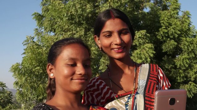Two women share mobile phone selfie video chat communication network  togetherness bonding in traditional Indian costumes in a rural village handheld two medium shot outdoor sun tree rural