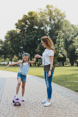 White sneakers. Young modern mother wearing white sneakers walking with daughter standing on skateboard