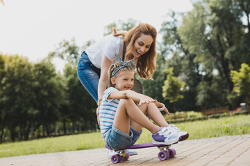 Young mother. Young loving mother watching her active girl sitting on skateboard in the park