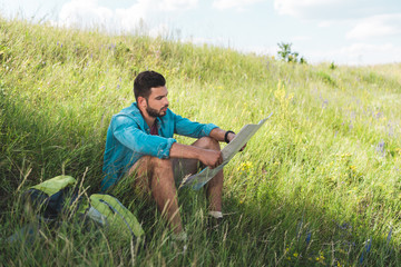 Naklejka na ściany i meble traveler sitting on green grass with backpack and looking at map