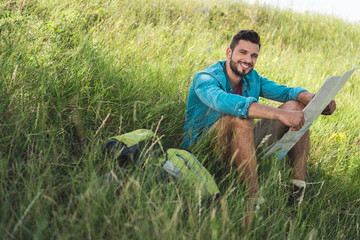 smiling traveler sitting on green grass with backpack and map