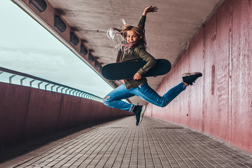 Happy schoolgirl with blonde hair dressed in trendy clothes jumps with a skateboard at a bridge...