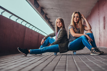 Mother and her daughter schoolgirl with blonde hair dressed in trendy clothes sitting together on a skateboard at a bridge footway.