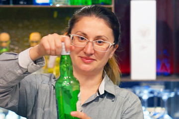 A bartender woman smiles and opens a bottle of whiskey for the client of the hotel bar. Shelves with bottles of alcohol in the background.