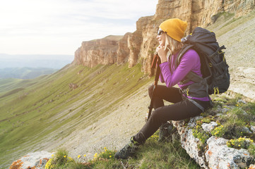 Smiling girl traveler in a yellow hat and a pair of sunglasses ssitting at the foot of epic rocks...