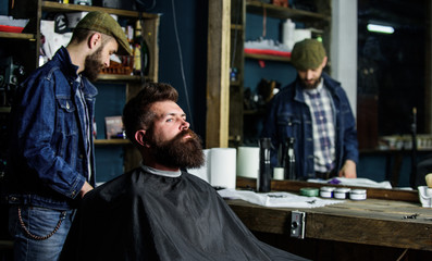 Hipster client getting haircut. Man with beard covered with black cape waiting while barber...