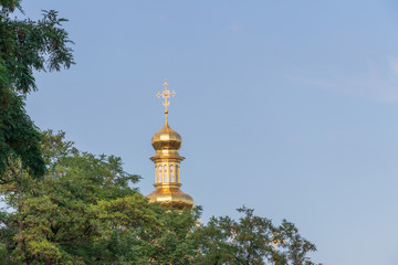 from behind the trees, you can see the top of the part of the golden dome of the church with a cross on the blue sky background