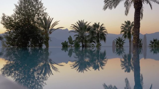Glassy and slightly shaking pool surface. Green palms and trees of different shapes and sizes are growing behind it. Beautiful misty mountains and a light blue and pink sky fill the background