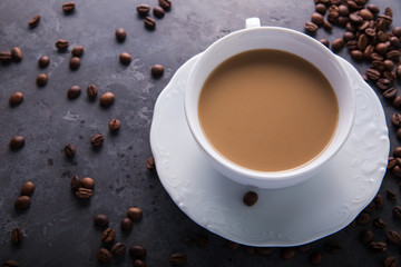  White cup of coffee with milk or tea with milk on dark grey beton background decorated with coffee beans. Copy space
