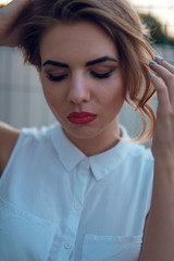 Close up portrait of tender blonde woman with beautiful make up, red lips posing at the camera outdoors in the evening. Urban background