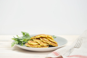 Small pancakes from a zucchini on a plate. Next to the pancakes are parsley. In the frame, cloth napkin and fork. Light wooden background. Close-up. Free space under the text.