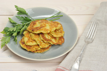 Small pancakes from a zucchini on a plate. Next to the pancakes are parsley. In the frame, cloth napkin and fork. Light wooden background. Close-up. View from above.