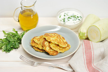 Small pancakes from a zucchini on a plate. Next to the dish there is a napkin, a fork, a bunch of parsley, a jug with olive oil and a cup with sour cream sauce. Light background. Close-up. 