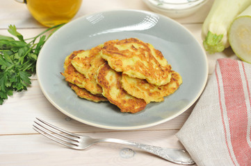 Pancakes from a vegetable marrow on a plate. Next to the plate is a napkin, a fork, fresh herbs and a zucchini. Light wooden background. Close-up. Macro photography.