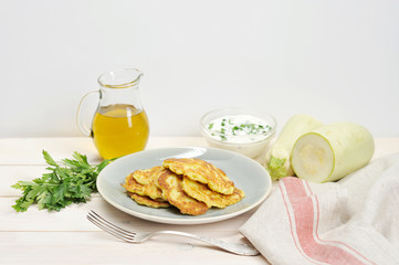 Pancakes from a vegetable marrow on a plate. Next to the plate is a napkin, a fork, a bunch of parsley, a jug of olive oil and a cup of sour cream. In the frame there is a fresh zucchini. 