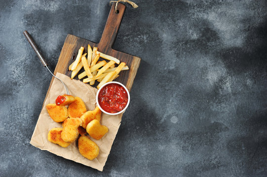 Chicken nagets on a wooden board. Rgdom with nuggets french fries and ketchup sauce. Dark background. Close-up. View from above. Vertical orientation of the frame. Free space for text.