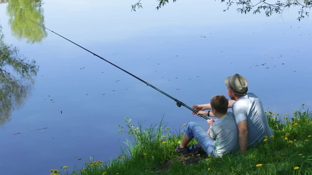 Back view of happy family on summer vacations concept. Father and son fishing together at river bank at scenic landscape background of fresh green grass and blue calm water.