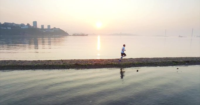 Aerial view of young man running faster and faster in the early morning along the spit. Ascending view of Tokarevsky lighthouse with sunrise, Vladivostok, Russia
