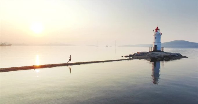 Beautiful aerial static view of man in shorts running fast along the Tokarevsky spit to the lighthouse. Fresh morning sea at amazing sunrise. Vladivostok, Russia
