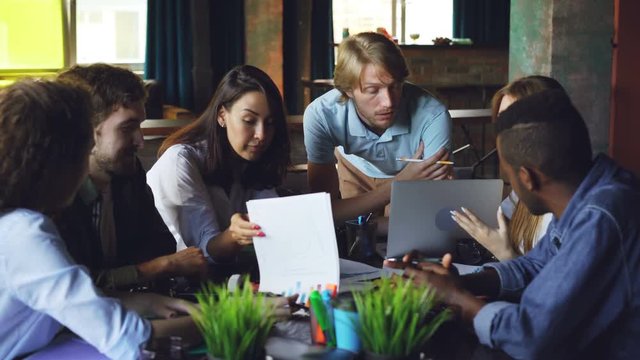 Young men and women colleagues are looking at papers lying on table and talking sharing ideas. Teamwork, modern office and company employees concept.