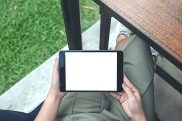 Top view mockup image of woman's hands holding black tablet pc with white blank screen while sitting in cafe with green nature background