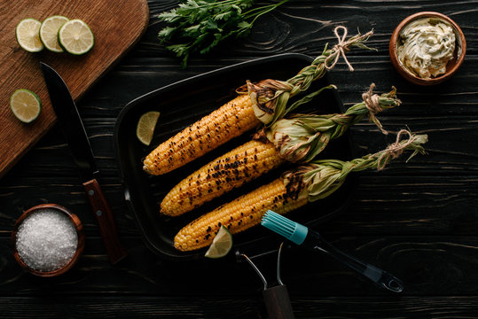top view of griddle pan with grilled corn and lime slices surrounded by salt and cream with parsley on wooden table