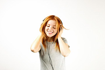 Young attractive natural redhead woman smiling dancing, singing along to her favorite song with large headphones. White background, copy space. Attractive female in casual outfit enjoying beloved tune