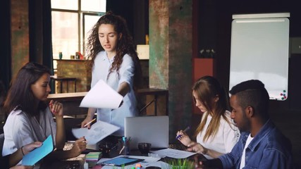Young woman team leader is giving papers with plans to group of young people Caucasian and African American and talking during meeting. Discussion and documents concept.
