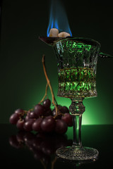 lead glass of absinthe with branch of grapes and burning sugar on spoon on reflective surface and dark green background - obrazy, fototapety, plakaty