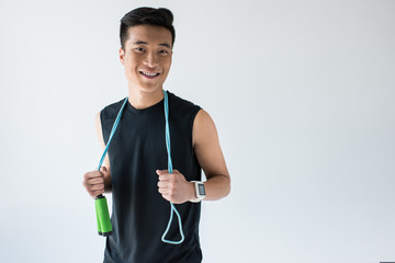 smiling asian sportsman with smartwatch holding jump rope isolated on grey background