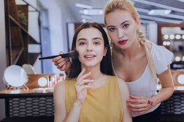 Service for client. Professional blonde-haired makeup artist servicing her beautiful client in beauty saloon