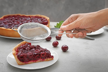 Woman sprinkling piece of tasty cherry pie with powdered sugar on table