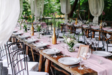 Wedding. Banquet. The festive table for guests, decorated with a composition of white and pink flowers and greenery, there are candles, served with crockery in wooden summer house.
