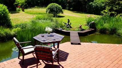 attraktiver naturbelassener Teich an rote Terrasse angrenzend mit Brücke und Tisch und Stühlen