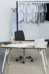 selective focus of t-shirts on hangers and working table with laptop and paintings in clothing design studio