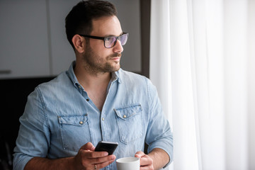 Happy handsome man drinking coffee in the morning texting on smartphone. Positive emotions