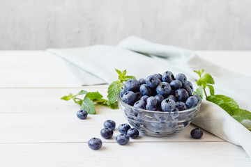 Fresh ripe blueberries in a glass bowl and mint on a light wooden table.