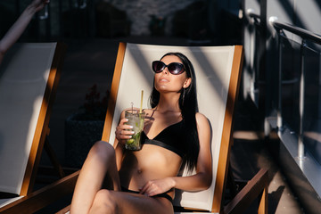 Outdoor summer lifestyle portrait of stylish sexy woman with perfect tanned fit body wearing trendy sunglasses drinking cocktail and enjoying pool party on luxury villa.