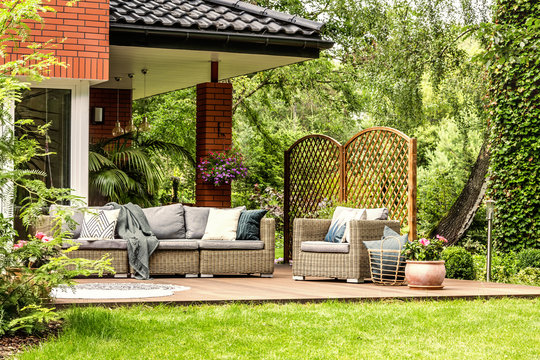Blanket thrown on garden sofa, basket with pillows standing next to armchair on wooden terrace