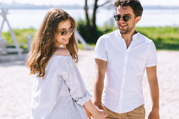 Obraz premium smiling couple in sunglasses holding hands at sandy city beach