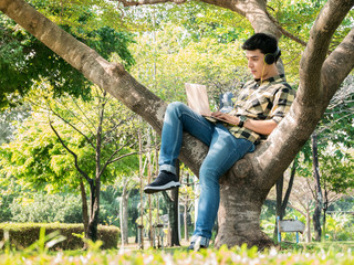 Young handsome man using laptop computer while listening to music with headphone sitting on the tree in the park.