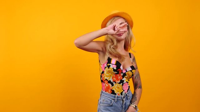 Happy sexy woman wearing sunglasses and a hat dances and smiles to the camera isolated on yellow orange background. Happines, travel, vacation and summer concept footage