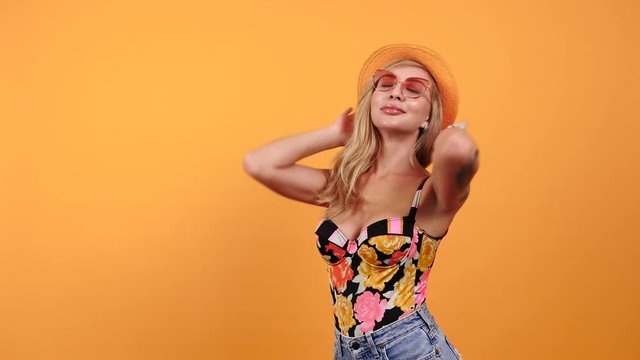 Hot sexy tanned woman wearing sunglasses and a hat smiles to the camera on yellow orange background. Copyspace available
